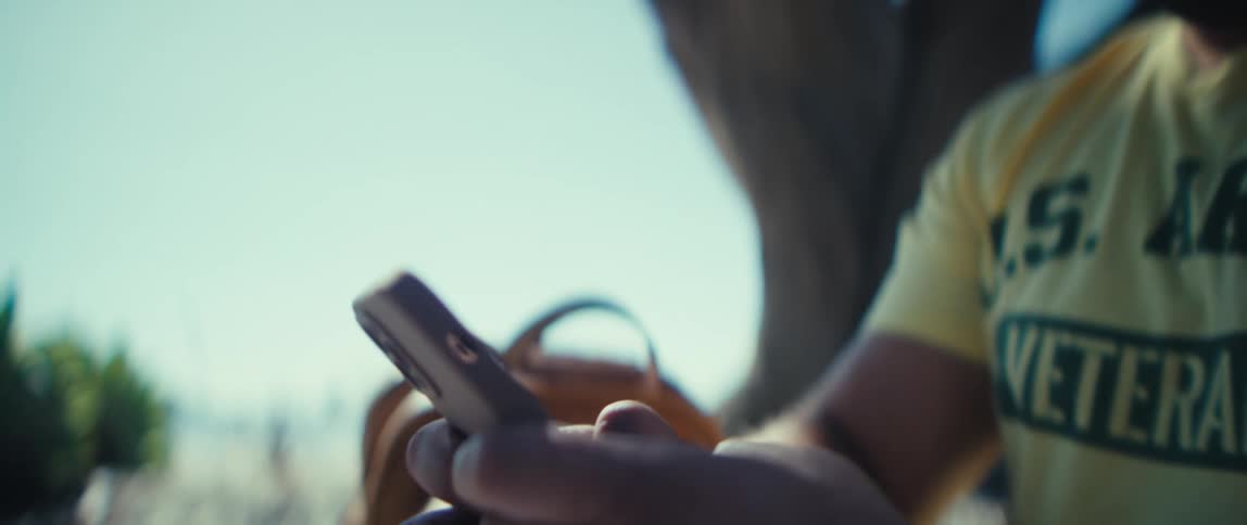 A man smiles as he sits under a tree and looks at his phone