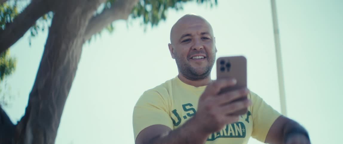 A man smiles and waves at his phone as he sits under a tree