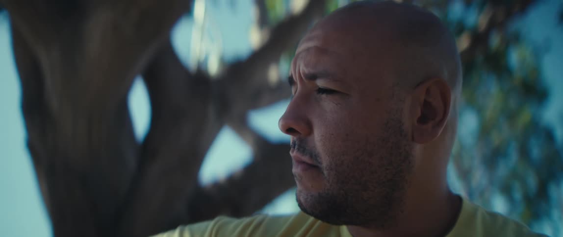 The man is seated outdoors partially shaded by the dark outoffocus branches of a tree visible in the background his gaze is directed off to the side as he appears to be lost in thought or observing something with intensity the lighting is soft and n