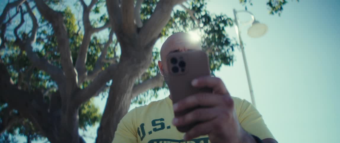 A bald man holding a his smart phone stares at its screen while outdoors