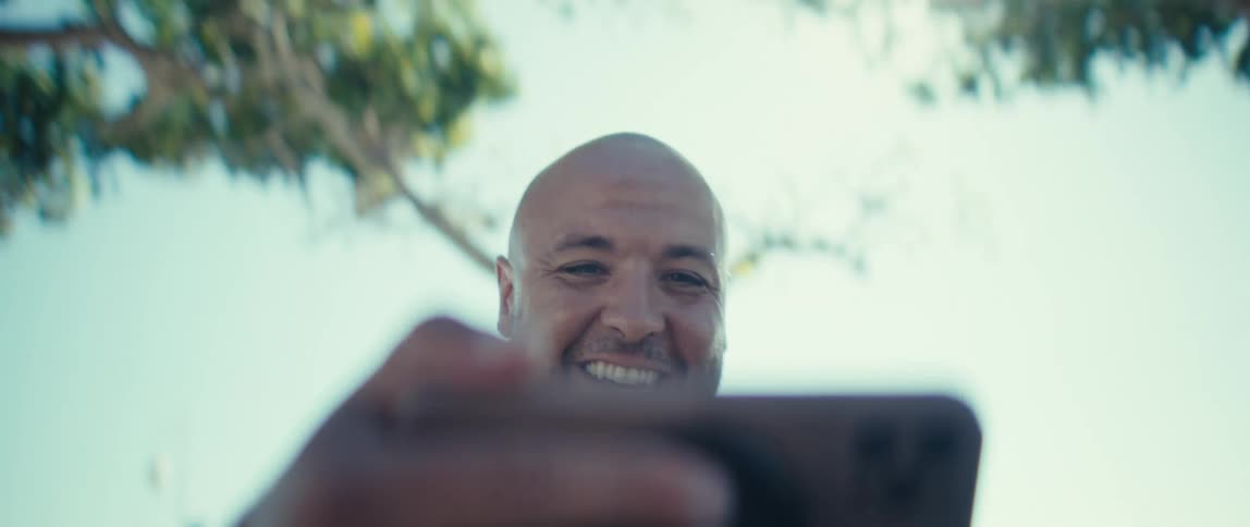 A bald man in a yellow shirt takes a selfie video with his phone in an outdoor setting on a very bright sunny day he smiles widely and pumps his fist in the air expressing excitement or joy