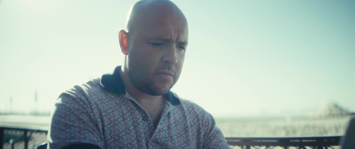 A man sitting near a metal fence separating him from a beach typing on a laptop with a serious stern look