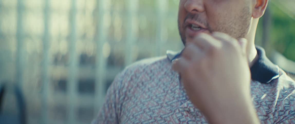 A man sitting at a table on a balcony speaks and gestures in front of an opened laptop during a video call