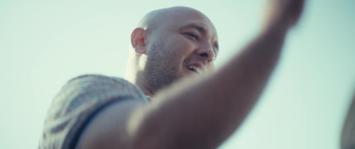 A mans face fills the frame captured from a low angle the bright sunlight from the sky which appears almost whitelight blue due to overexposure backlights his head and shoulders he is smiling gently and his eyes seem to be following something or so
