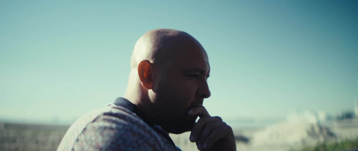 The man is shown from the side standing outdoors on a bright day with a pale blue sky and hazy outoffocus background he has a serious expression and his fingers touch his chin or mouth suggesting he is mulling over an idea or focused on a distant o