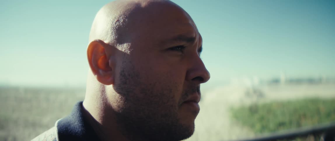 Young bald man on a balcony next to a beach typing on a laptop computer on top of a table outdoors during day time