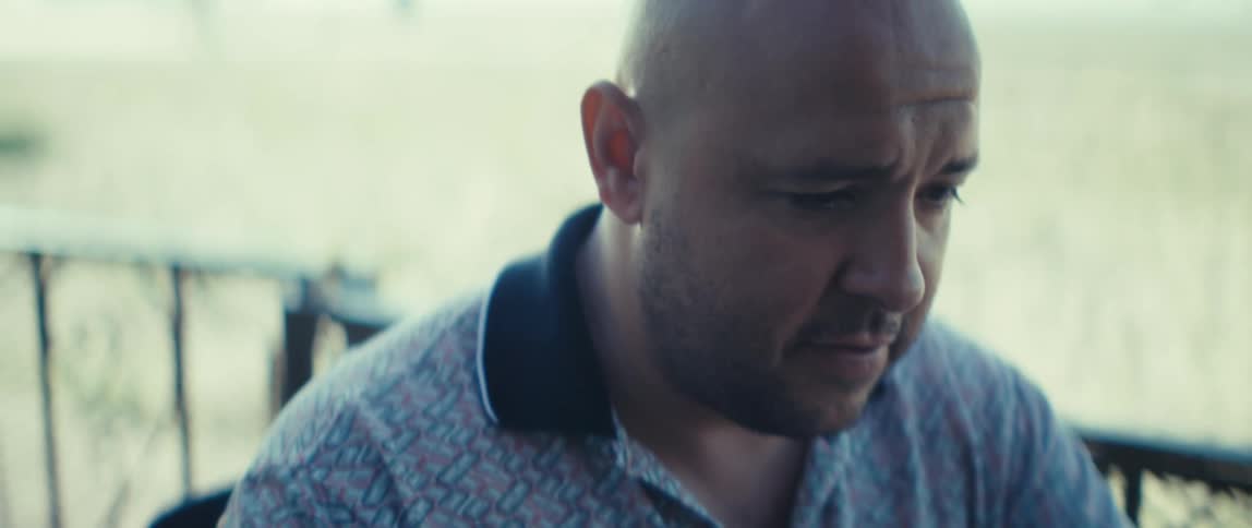 Young bald man wearing a polo shirt sitting on a balcony next to a beach talking and using a laptop computer on top of a table outdoors during day time