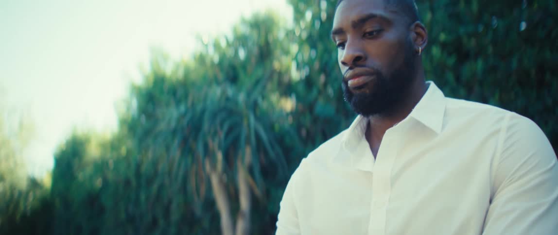 A closeup shot of a man wearing a crisp white shirt focuses on his contemplative expression as he looks down and then slightly to his side the camera then tracks up slightly showing him holding a tablet and stylus while the bright green foliage of the