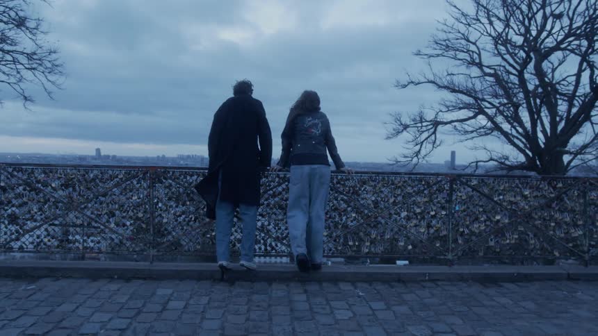 Two people leaning on a padlocked fence overlook an urban skyline under a cloudy sky
