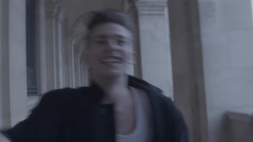 The clip is a shaky closeup shot that follows a young man as he runs looking overjoyed through a long colonnaded stone corridor the background consists of repeating arches and pillars creating a sense of depth the muted lighting and the filmsupply