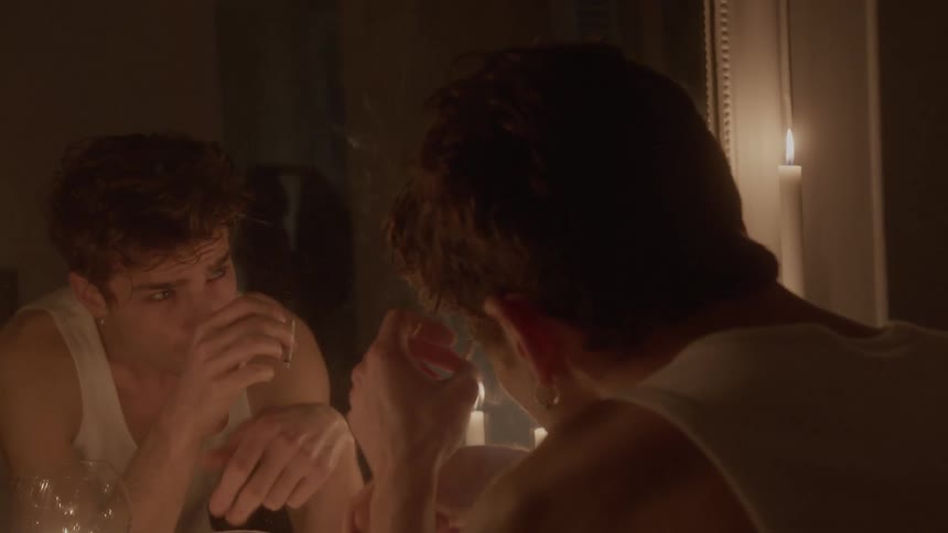 A young man sitting in front of a mirror smokes a cigarette with a sad expression as he watches the reflection of a woman getting dressed behind him