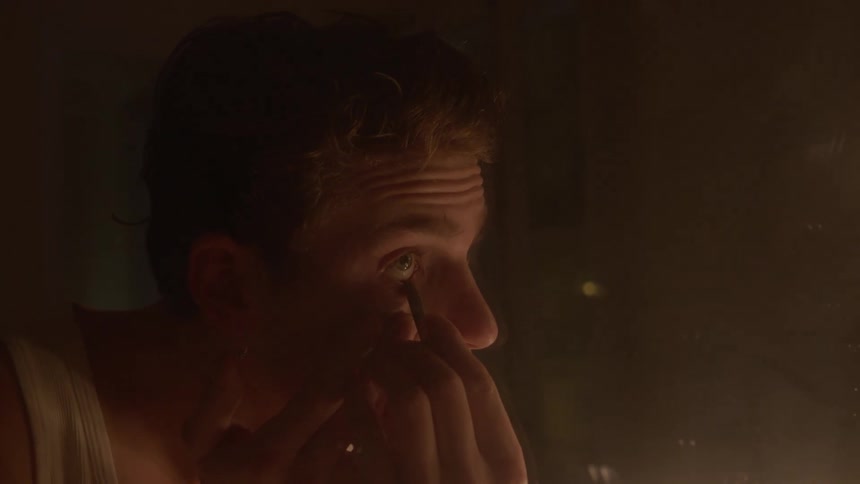A young man draws his eyebrows in front of a mirror with a candle lighting the dark room