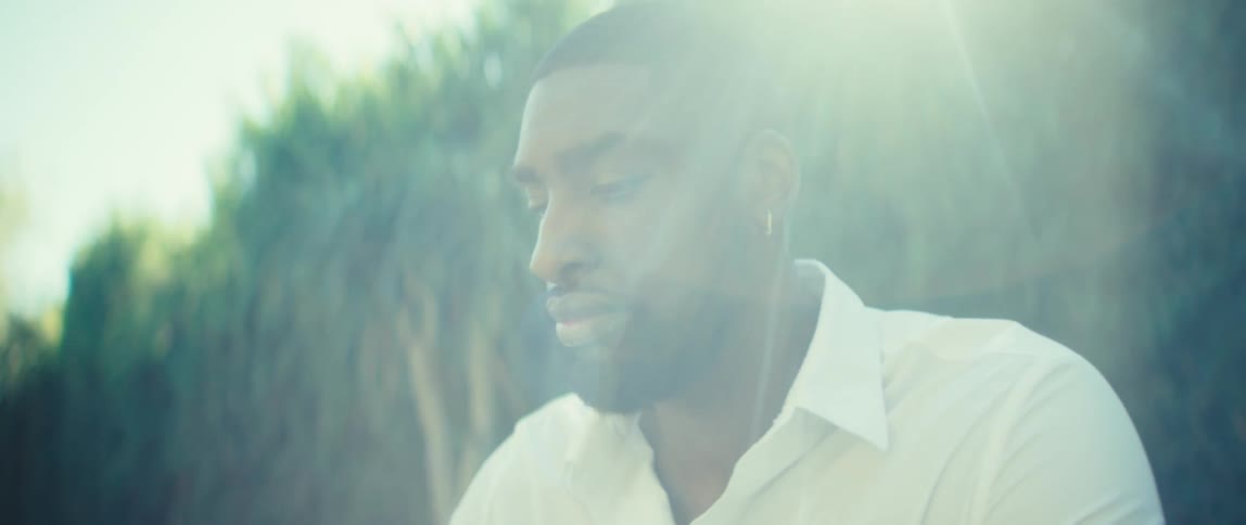 A man wearing a white collared shirt is seated outdoors initially looking down in concentration as he uses a stylus on a tablet held in his hands he then lifts his head and looks off to the side with a serious thoughtful expression while a bright lens