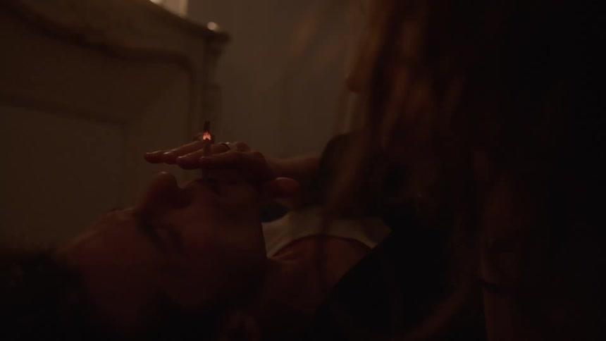 A man lies back with eyes closed in warm candlelight relaxed as he smokes a cigarette a woman close beside him holds to his mouth