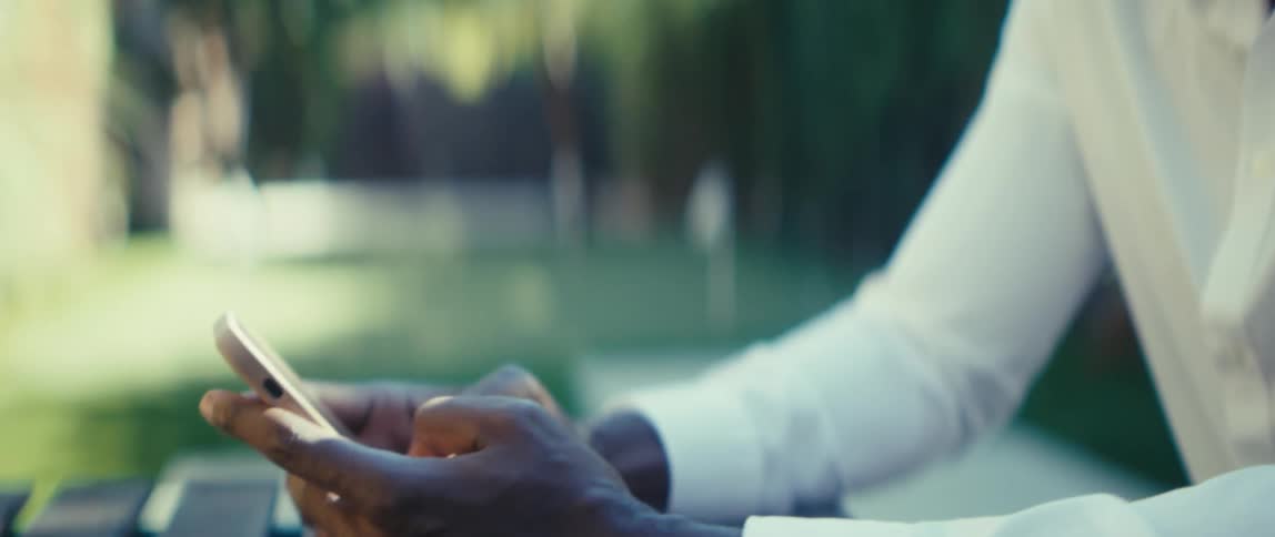 Person in a white shirt holds a smartphone while seated at a wooden table outdoors with blurred greenery in the background