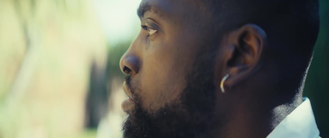 A young man wearing earrings sits outdoors with facial hair at midday