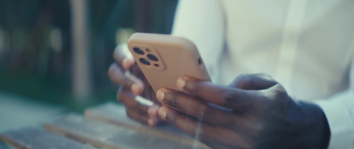 A young man sitting outdoors behind a coffee table using his mobile phone at midday