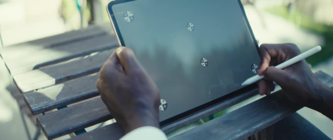 A young man sitting on a chair outdoors with a coffee table in front of him using a tablet with a pen on a sunny day