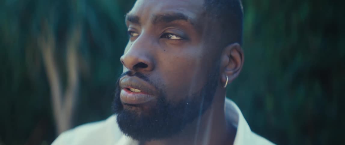 A young black man wearing earrings and a milkcolored shirt sits outdoors in a backyard at midday