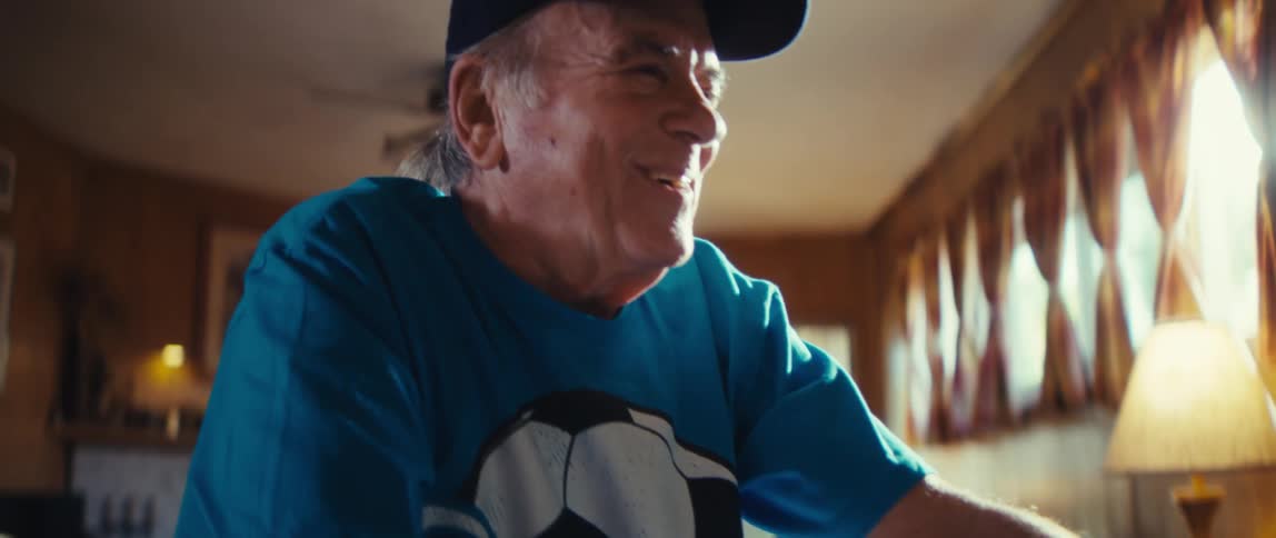 A n elderly in a teal shirt holds a tiny soccer ball at a sunlit table as he smiles and cheers