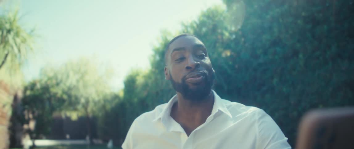 Man in a white shirt holds a smartphone in an outdoor area with tall greenery and blue sky in the background