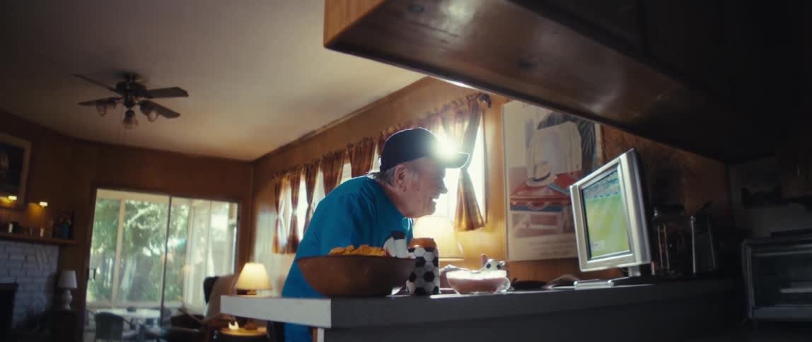 An elderly man in a blue shirt and a baseball cap is shown in an indoor setting leaning over a counter to intensely watch a soccer game on a computer monitor he smiles widely and enthusiastically around the foursecond mark clearly enjoying the game a