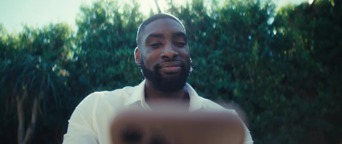 A black man in a white shirt holds a phone at arms length outdoors looking calm and confident with greenery behind him