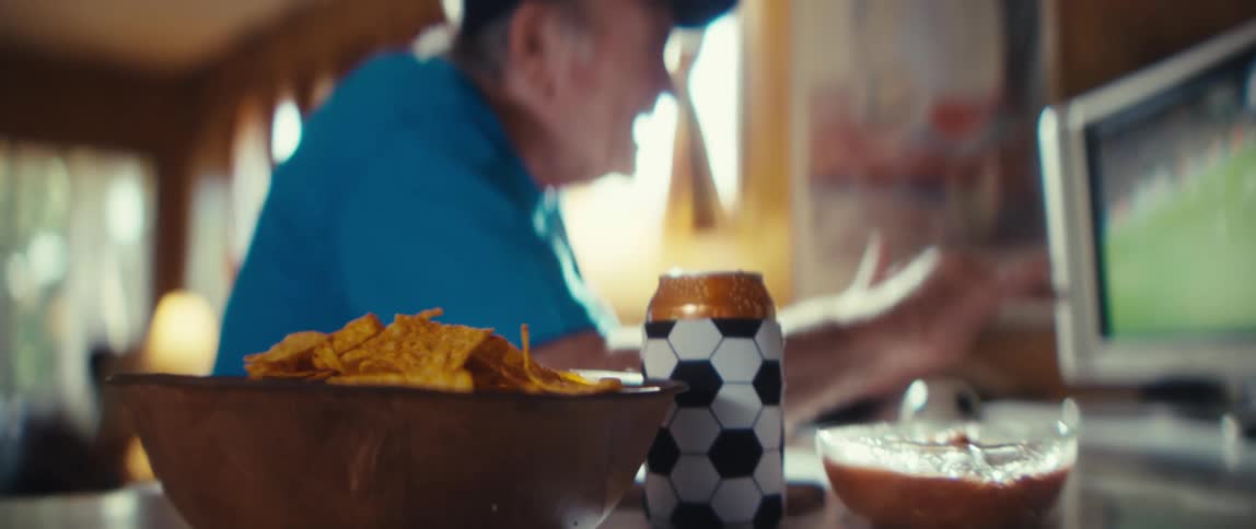 An elderly man sits in his living room watching a soccer match on tv in excitement wearing a blue tshirt and a hat with a bowl of chips and a can of beer on the table beside him