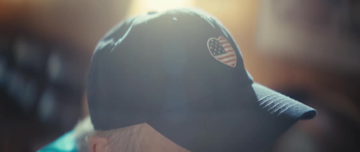 An elderly man sitting indoors wearing a hat with the us flag on it enjoying his time inside