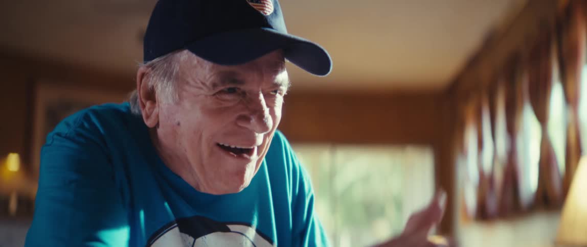 A smiling older man in a navy baseball cap gives an enthusiastic thumbs up while looking at a tv screen