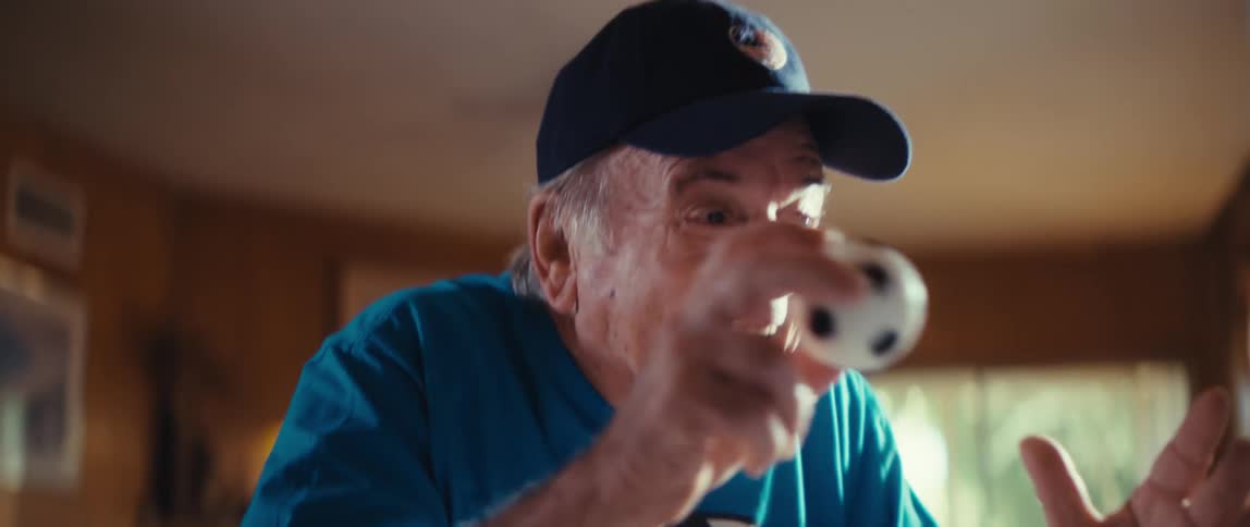 Elderly man in a blue cap and graphic shirt smiles while looking at a screen in a woodpaneled indoor setting