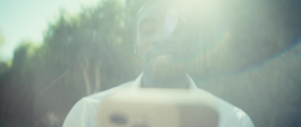 A man with a neatly trimmed beard and dark hair is seen outdoors in a closeup shot initially looking seriously at his phone held in the foreground after a brief moment a big smile spreads across his face as he looks up with bright sunlight and a lens