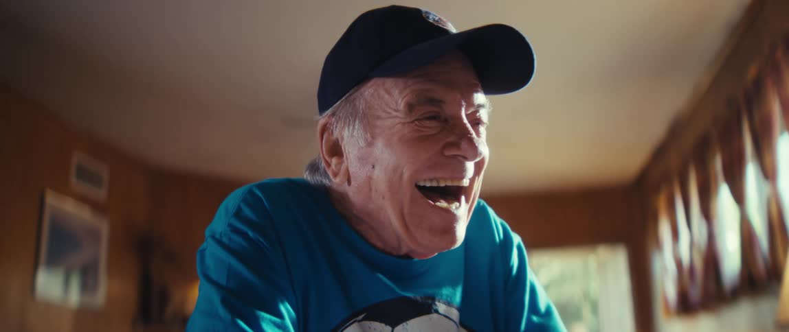 An elderly man looks excited as he bounces a small ball on a desk in his living room smiling and laughing while wearing a blue tshirt and a hat