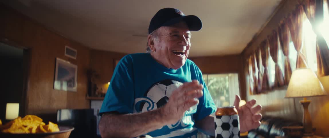 An elderly man wearing a blue shirt that says soccer grandpa and a baseball cap is initially leaning forward watching a screen with a serious intense expression suddenly he shouts in excitement throwing his arms up in the air in a triumphant celebr