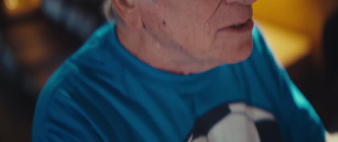 The video starts with a closeup on an older man wearing a dark baseball cap featuring a heartshaped american flag patch on the front as the camera pans down it reveals the mans excited openmouthed expression and his blue tshirt with a large soccer