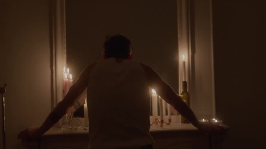 A man seen from the back in silhouette stands in a dimly lit room leaning on a mantelpiece or table with his arms spread slightly the only light comes from multiple tall flickering candles arranged around a large mirror or window casting an intense