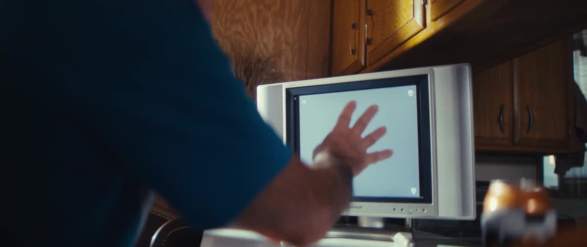 An elderly man watches a football game at midday holding a small ball and wearing a blue tshirt and a hat excited as he looks at the tv