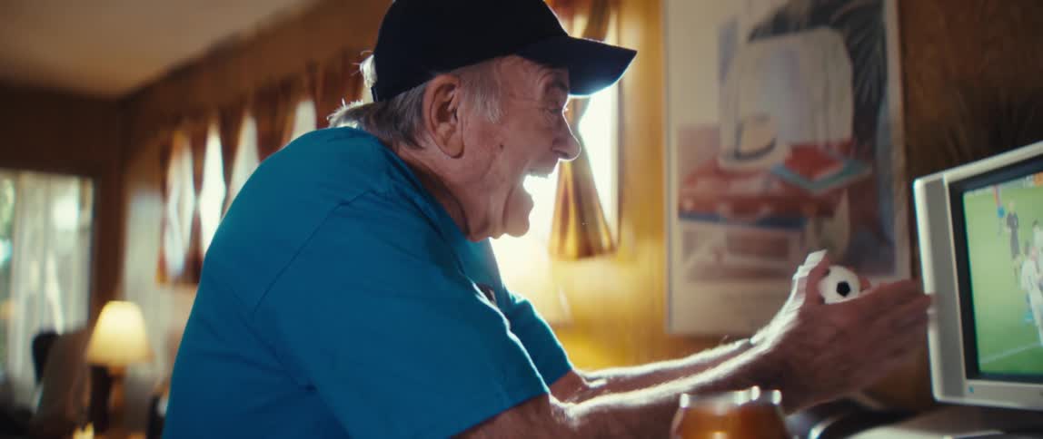 An elderly man sits in his living room watching a soccer match on tv in excitement wearing a blue tshirt and a hat with a bowl of chips and a can of beer on the table beside him