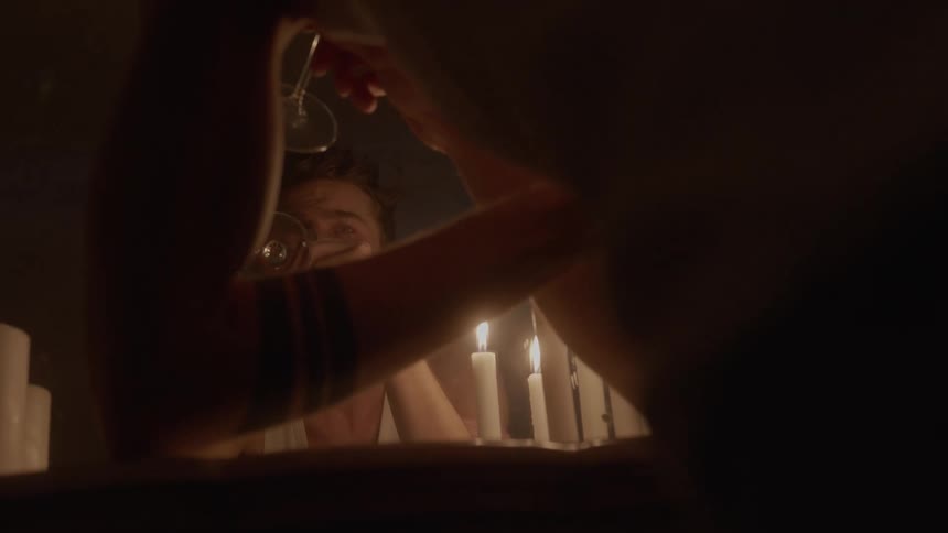 The camera captures a closeup of a mans face reflected in a mirror dimly lit by the candles positioned on a surface between him and the camera he stares intently at his reflection then slowly raises a wine glass to his lips taking a slow sip