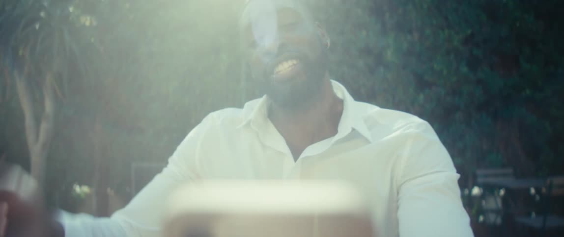 A smiling man in a white buttonup reaches outward and talks in his phone in hazy sunlight in a leafy backyard