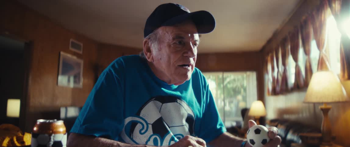Elderly man in a soccerthemed outfit engages with a screen while holding a ball and enjoying snacks