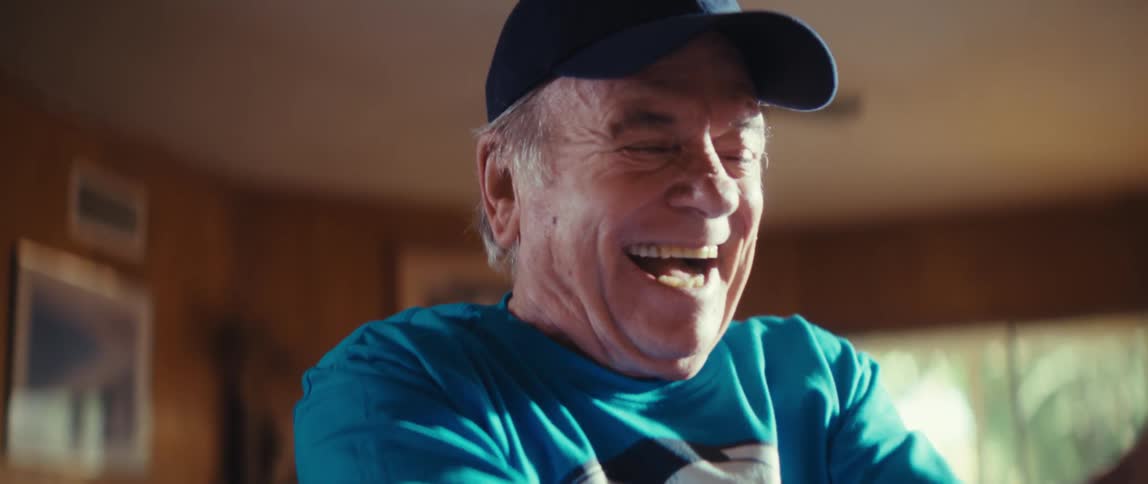 An elderly man wearing a soccer fan t shirt and a cap with a heart shaped american flag and holding a small soccer ball stands up to clap and cheer with a large smile on his face