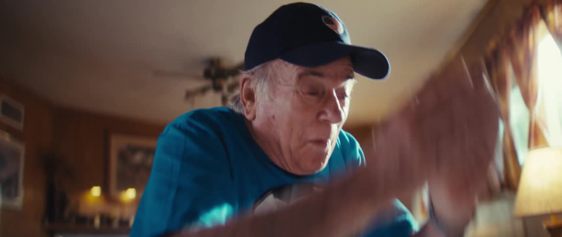 An elderly man wearing a hat plays indoors bouncing a small ball and having fun at midday