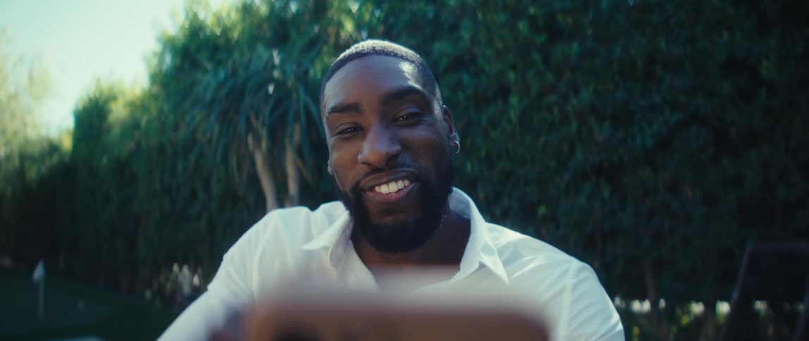A man in a backyard surrounded by trees waving and smiling in a video call on a smart phone