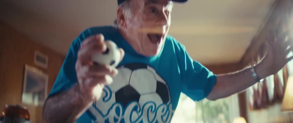 Elderly man in a dark cap and soccer shirt holds a small ball while looking at a screen in a woodpaneled indoor setting