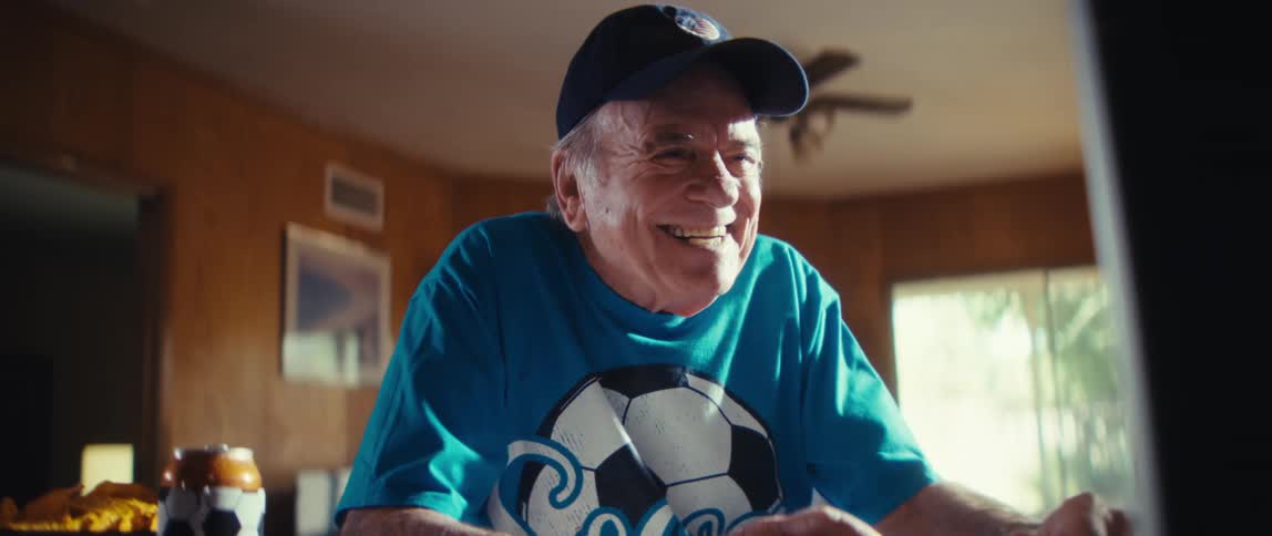 An elderly man wearing a soccer fan t shirt a cap with an american flag and holding a small soccer ball moves and smiles excitedly as he celebrates while watching a game on television