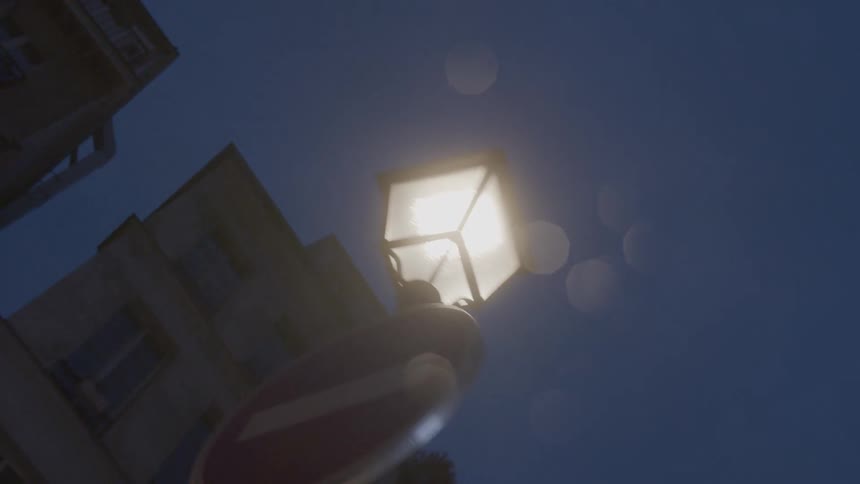 A glowing streetlamp at dusk shines above a noentry sign seen from a low angle with soft bokeh and building edges in the background