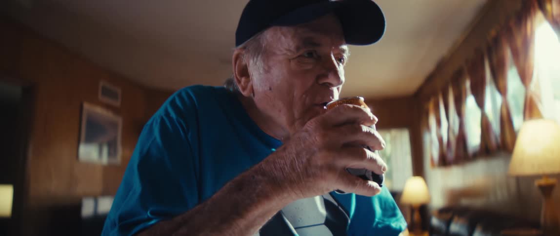 An elderly man wearing a soccer fan t shirt a cap with an american flagwith a small soccer ball to the right of his hand cracks open a can with the image of a soccer ball on it drinks from the can and smiles
