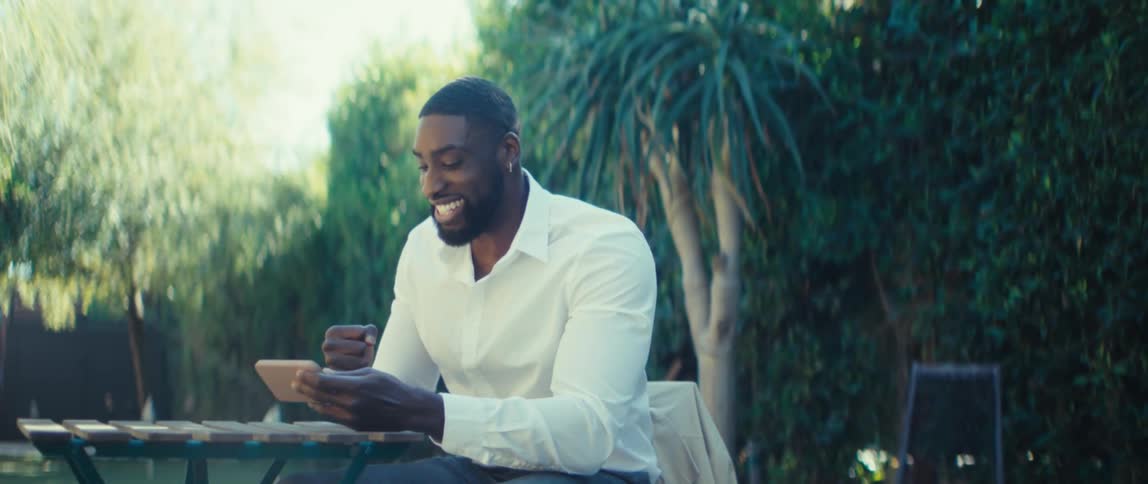 A young black man sitting in a backyard watches content on his mobile device looking excited while wearing a longsleeve shirt and surrounded by trees on a sunny day