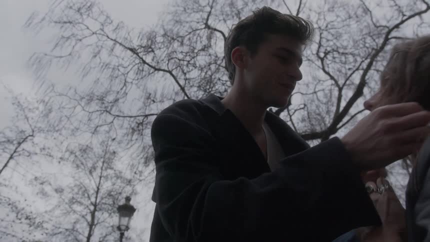 A young man and woman are seen in a close intimate moment outdoors under a somber overcast sky with bare tree branches above them the man initially has his arm around the womans head as she looks down then he gently lifts her chin to meet his gaze be